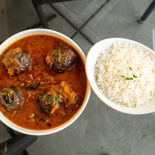 Eggplant curry in a white bowl next to a bowl of white rice, garnished with herbs.