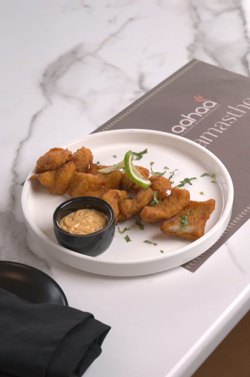 Crispy fried fish pakora with a side of dipping sauce and lime on a white plate.