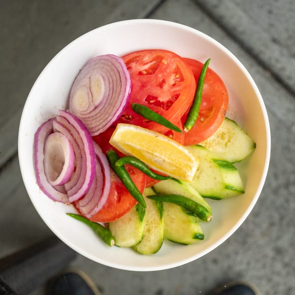A white bowl filled with sliced red onions, tomatoes, cucumbers, green chilies, and a lemon wedge.