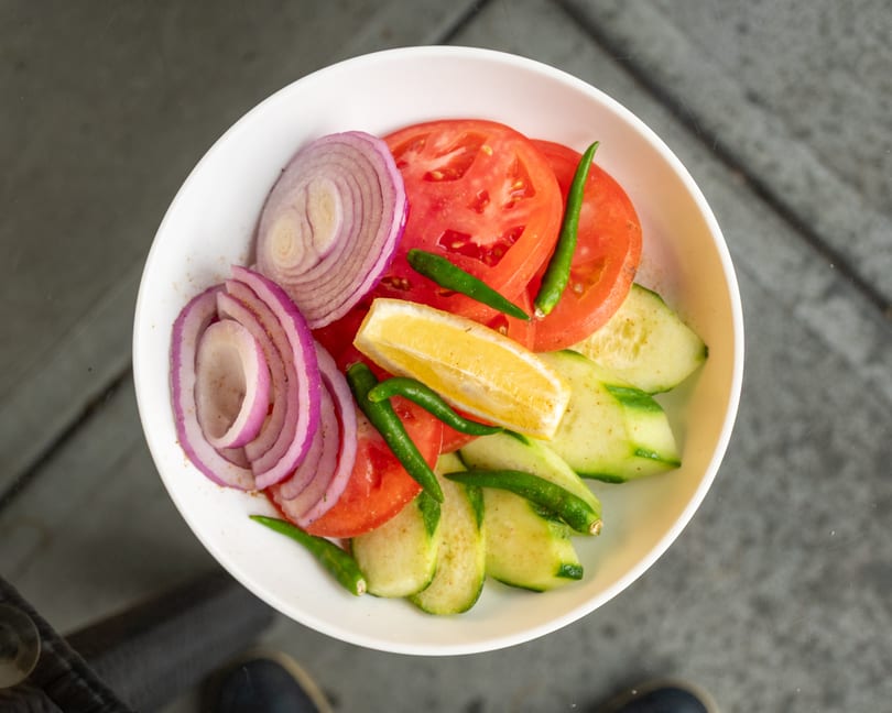 A white bowl filled with sliced red onions, tomatoes, cucumbers, green chilies, and a lemon wedge.