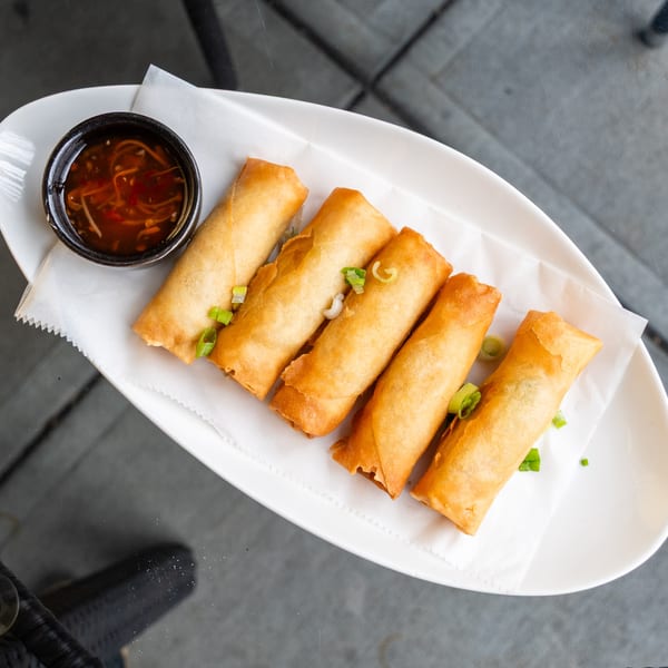 Five crispy golden spring rolls with green onions and a side of dipping sauce on a white plate.