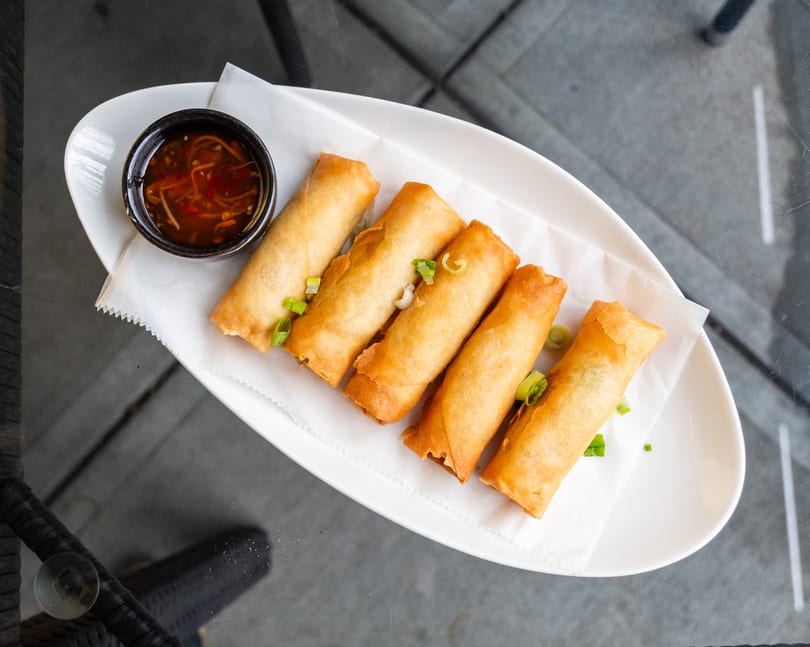 Five crispy golden spring rolls with green onions and a side of dipping sauce on a white plate.