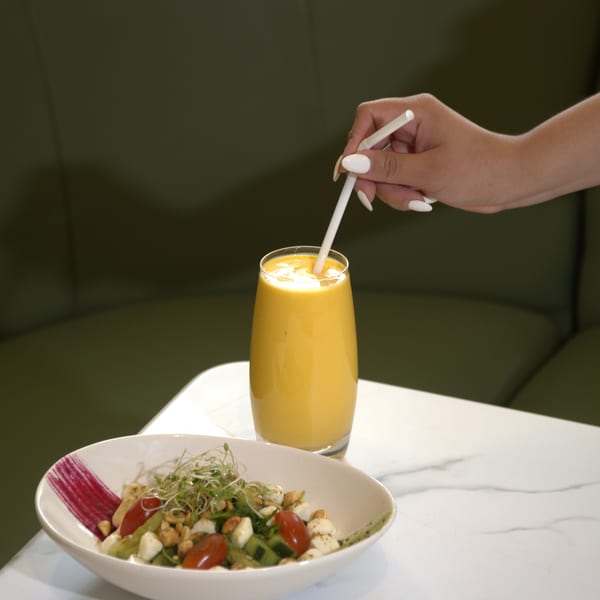 A hand stirs a yellow smoothie next to a fresh salad with cherry tomatoes and sprouts.