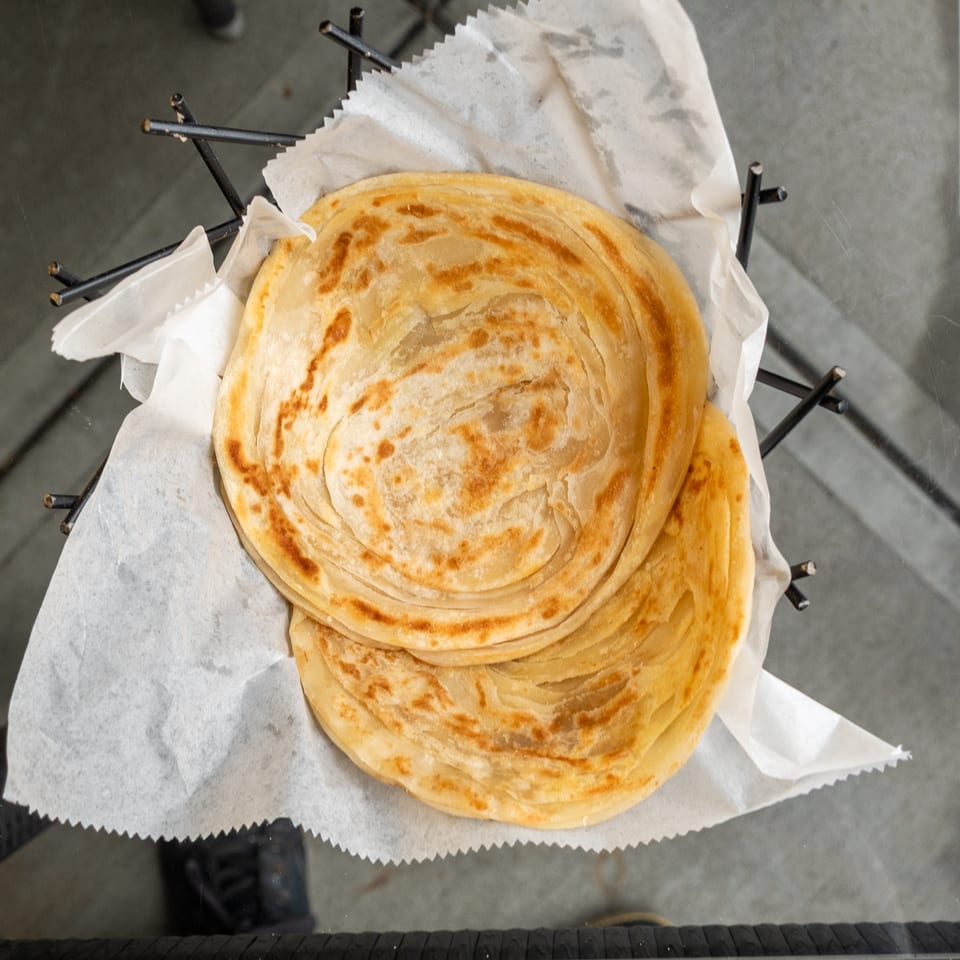 Two golden-brown, flaky paratha flatbreads served in a black wire basket lined with white paper.
