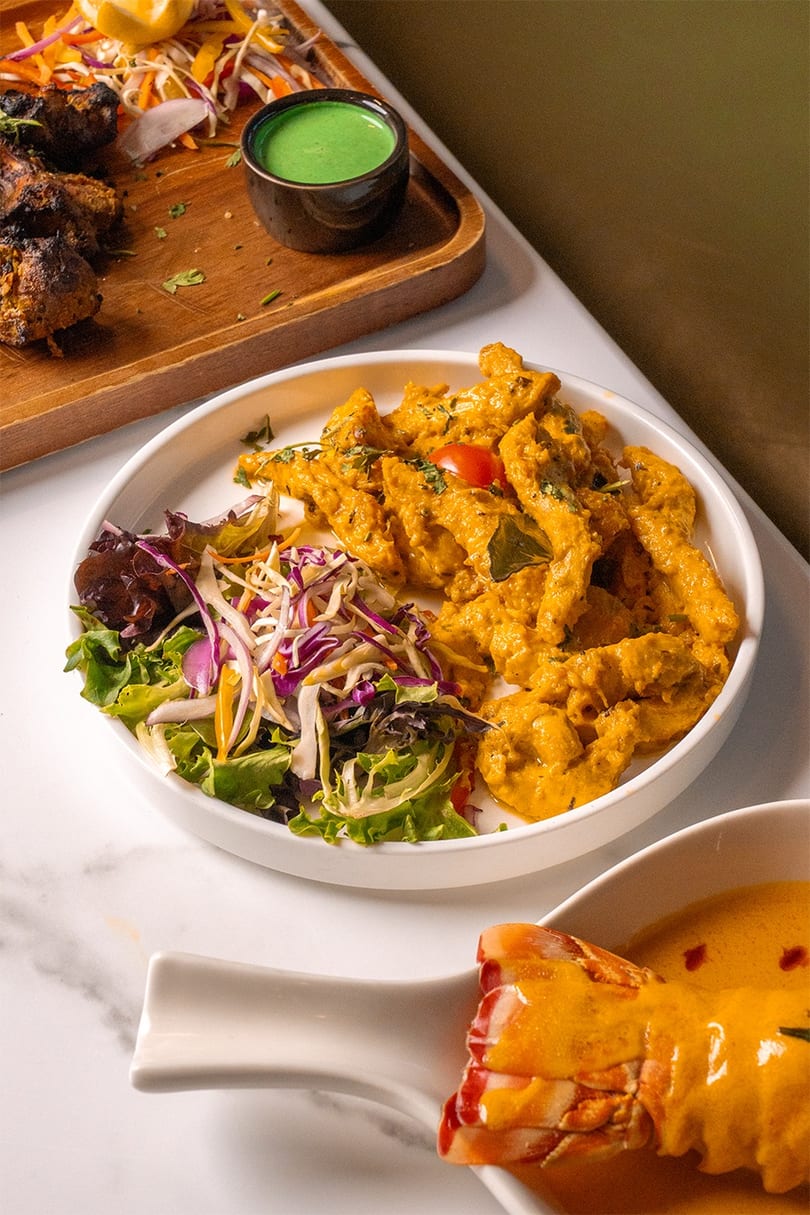 A white bowl of chicken in orange sauce with a side salad, next to a lobster tail.