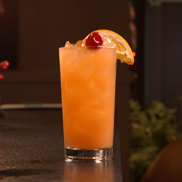 A vibrant orange cocktail with ice, garnished with a cherry and an orange slice on a dark bar.