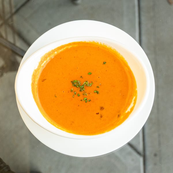 Overhead view of a white bowl of creamy orange tomato soup garnished with green herbs on a glass table.