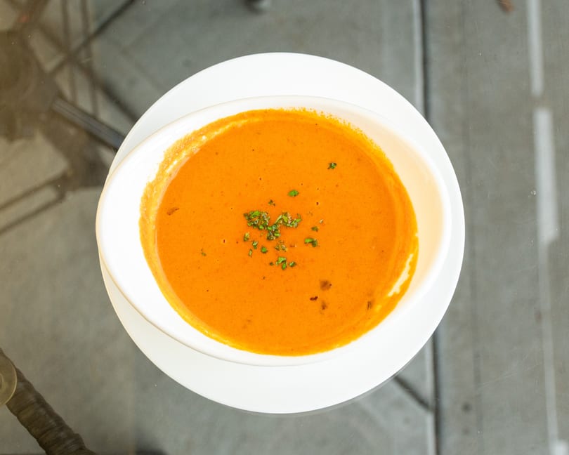 Overhead view of a white bowl of creamy orange tomato soup garnished with green herbs on a glass table.