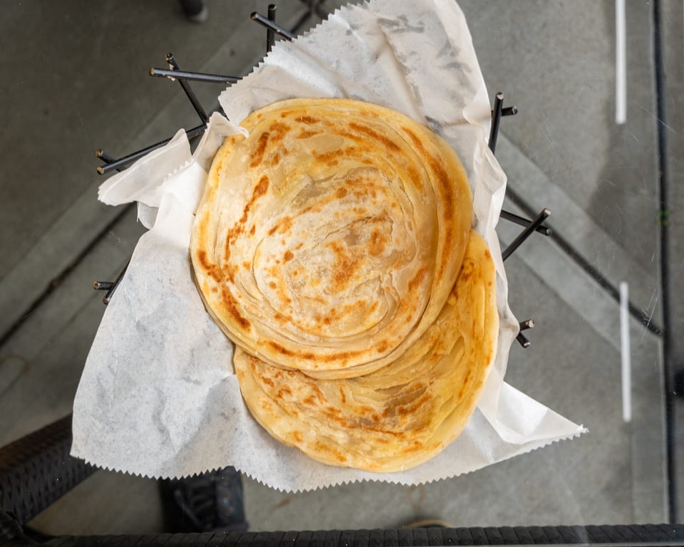 Two golden-brown, flaky paratha flatbreads served in a black wire basket lined with white paper.