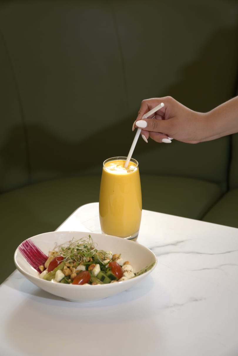 A hand stirs a yellow smoothie next to a fresh salad with cherry tomatoes and sprouts.
