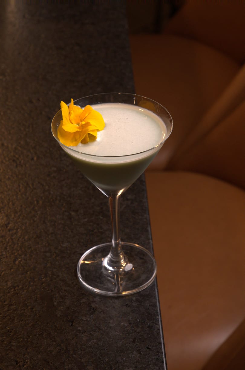 A green cocktail with a foamy top, garnished with a bright yellow flower, on a dark bar.