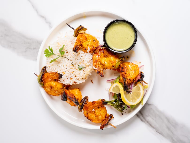 Grilled shrimp skewers with rice, a side salad, lemon, and green dipping sauce on a white plate.