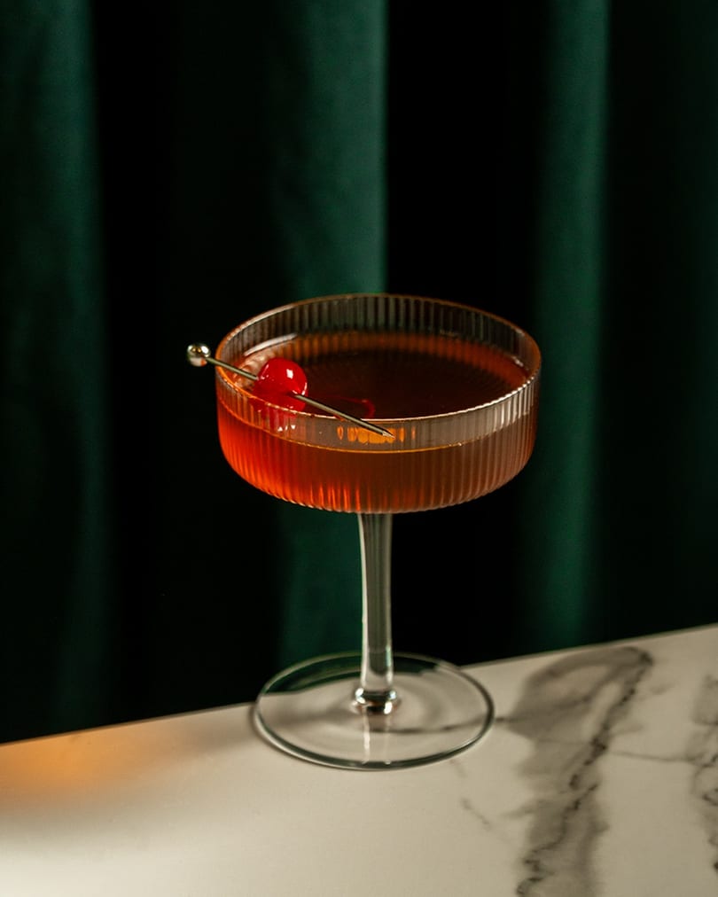 A Manhattan cocktail in a ribbed glass, garnished with a cherry, on a marble table.