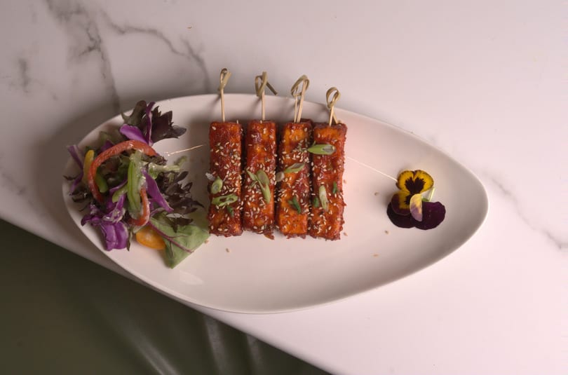 Four skewers of crispy tofu with sesame seeds and scallions, served with a side salad and edible flower.
