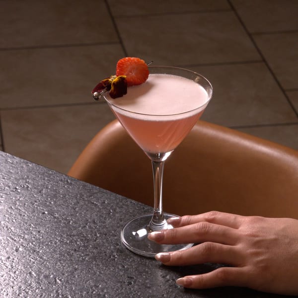 A pink martini cocktail garnished with a strawberry and flower, with a hand resting nearby.