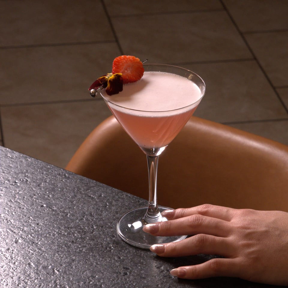 A pink martini cocktail garnished with a strawberry and flower, with a hand resting nearby.