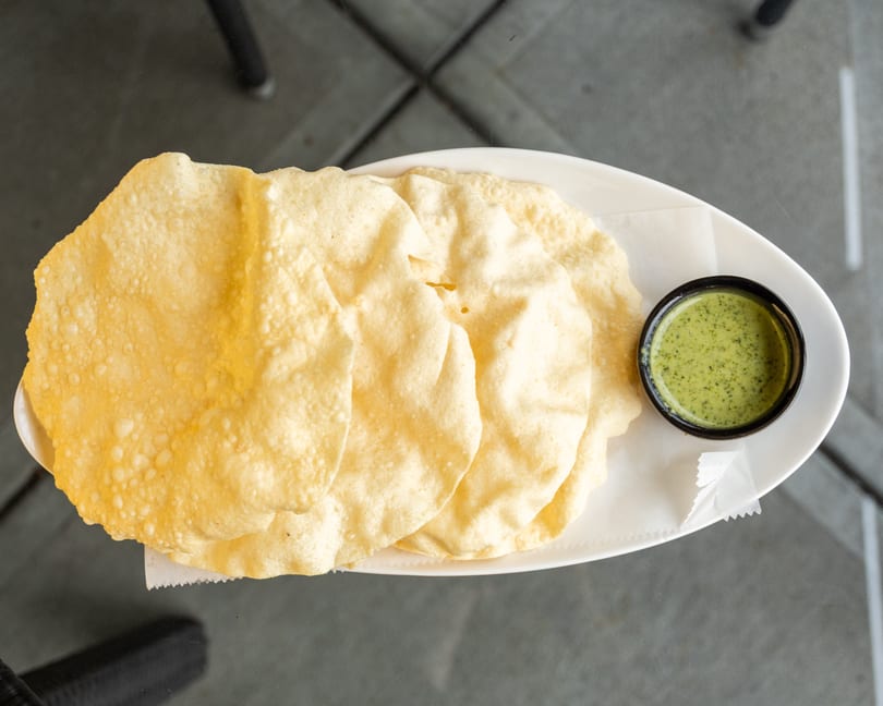 A white oval plate holds several crispy papadum with a small bowl of green chutney.