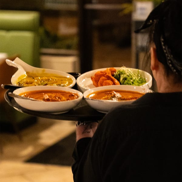 Server carrying a tray with four bowls of colorful Indian dishes through a restaurant.
