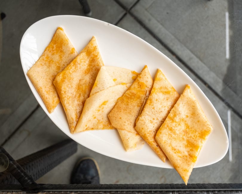 A white platter holds several triangular, golden-brown crepes, possibly filled with cheese, on a glass table.