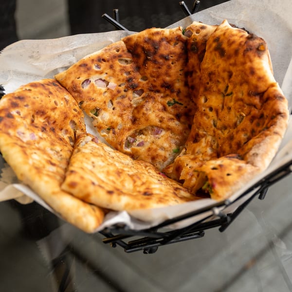 A basket of golden-brown naan bread, speckled with red onion and herbs, sits on a glass table.